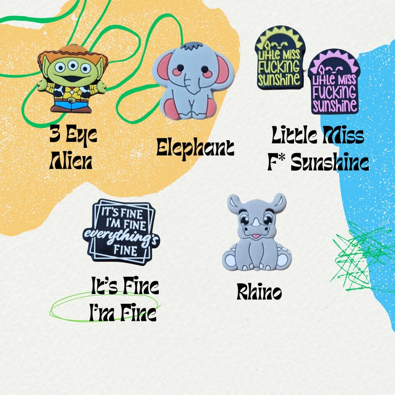 May include: A set of nine colorful shoe charms featuring cartoon characters and phrases. The charms include a three-eyed alien, an elephant, a rhino, and a charm with the phrase "It's Fine I'm Fine Everything's Fine". There are also two charms with the phrase "Little Miss F* Sunshine".