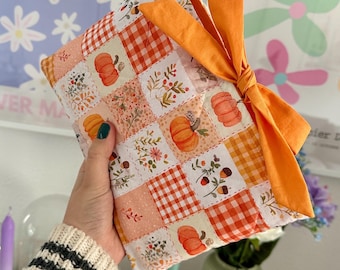 May include: A quilted fabric pouch featuring a patchwork pattern of orange pumpkins, acorns, floral sprigs, and orange gingham squares. The pouch is finished with a solid orange fabric bow tied on the side, creating a soft, textured appearance.
