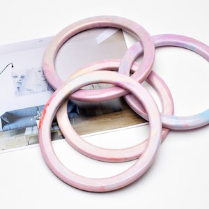 May include: Four pink marbled bangles are interlocked, with a photograph in the background. The bangles have a smooth, glossy finish and a mix of pink, white, and pastel colors. The photograph shows a modern interior with a sofa and artwork.