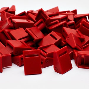 Może przedstawiać: Stos jaskrawoczerwonych plastikowych klamer, każda o prostokątnym kształcie i szczelinie na pasek. Klamry są rozrzucone na białej powierzchni, tworząc atrakcyjną wizualnie fakturę. Prawdopodobnie są używane do różnych projektów.