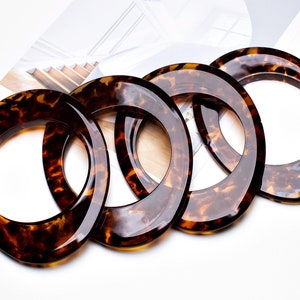 Puede incluir: Cuatro asas ovaladas para bolsos con un patrón de carey. Las asas están hechas de un material brillante y translúcido, con una combinación de colores marrón y ámbar. Están dispuestas en diagonal sobre una superficie blanca.