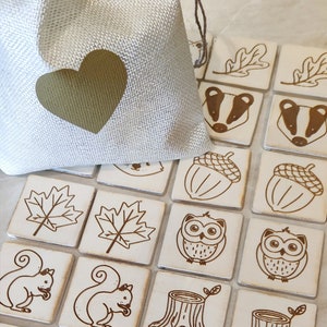 Könnte beinhalten: Ein Satz aus Holzfliesen mit verschiedenen Waldtieren und -gegenständen, darunter Eulen, Eichhörnchen, Hirsche, Eicheln, Blätter und Baumstümpfe. Die Fliesen sind in einem Gittermuster angeordnet und von einer weißen Oberfläche umgeben. Ein kleiner Jutesack mit einem goldenen Herzen befindet sich in der oberen linken Ecke des Bildes.