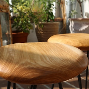 Könnte beinhalten: Zwei Holzstühle mit hellbraunen, ovalen Sitzen und schwarzen Metallbeinen. Die Hocker stehen im Freien, wobei Sonnenlicht die Holzmaserung hervorhebt. Der Hintergrund umfasst Topfpflanzen und einen rustikalen Spiegel.