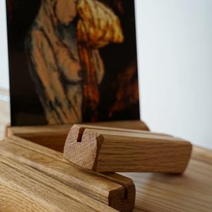 Könnte beinhalten: Holz-Bilderhalter mit einem Schlitz zur Aufnahme eines Fotos oder Kunstwerks. Die Halter sind aus hellem Holz gefertigt und rechteckig mit abgeschrägter Kante. Im Hintergrund befindet sich ein Kunstwerk einer Frau.