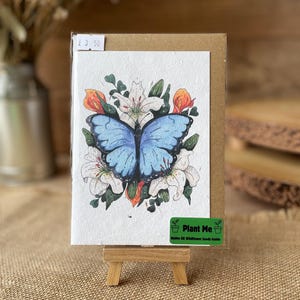 May include: A watercolour greeting card depicting a blue butterfly surrounded by white lilies and orange flowers. The card is displayed on a small wooden easel. A green sticker on the card reads "Plant Me" with text below.