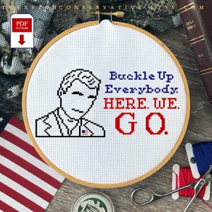May include: A cross-stitch embroidery hoop featuring a pixelated image of a person with the text "Buckle Up Everybody. HERE. WE. GO." in blue and red. The design includes an American flag and is set against a background with an American flag and a camouflage pattern.