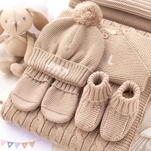 May include: A beige knitted baby outfit set with a hat, mittens, booties, and a sweater. The hat has a pom-pom on top and the sweater has the name "LENNON" embroidered on it.