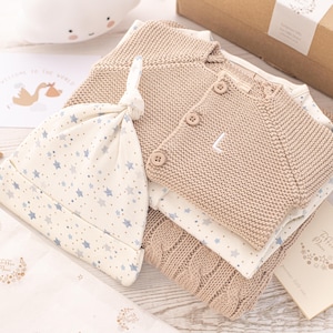 May include: A baby gift set with a beige knitted cardigan, a beige knitted hat, and a white onesie with blue stars. The set is packaged in a brown box with a white ribbon.