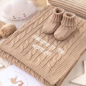 May include: A brown cable knit baby blanket with the text "Welcome to the World [Name]" embroidered on it. Two small brown knitted booties are on top of the blanket.