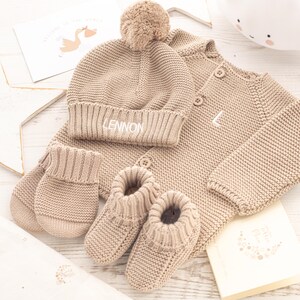 May include: A beige knitted baby outfit set with a hat, booties, and mittens. The hat has a pom-pom on top and the name "LENNON" embroidered on the brim.