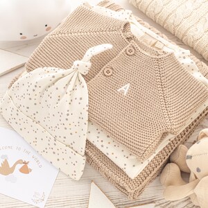 May include: A beige knitted cardigan with a white letter A embroidered on the front, a white and beige hat with a star pattern, and a beige blanket with a star pattern. The text "Welcome to the world" is visible on a card.