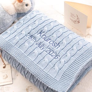 May include: A light blue knitted baby blanket featuring a cable knit pattern and embroidered text that reads Kourosh 28th July 2026. The blanket is accompanied by a small branded card and a plush toy visible in the upper corner of the frame.