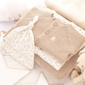 May include: A beige knitted baby cardigan with a white letter A embroidered on the front, a beige and white star patterned baby hat, and a beige blanket with a star pattern.
