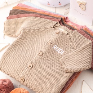 May include: A beige knitted baby cardigan with white buttons and the name "Rudi" embroidered on the front.