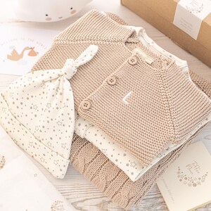 May include: A beige knitted cardigan, a white onesie with a star pattern, and a white knitted hat with a star pattern. The items are arranged on a white wooden surface. The text "Welcome to the world, little one" is visible on a card.