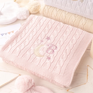 May include: A pink cable-knit baby blanket with an embroidered crescent moon, stars, and the letter "B". The blanket is folded and stacked with other blankets in cream and white. A matching hat with a pom-pom is in the foreground.