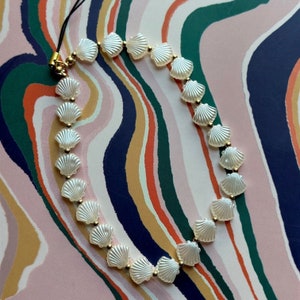 Può includere: Una collana di conchiglie bianche con accenti dorati. La collana è composta da più piccole perle di conchiglia e infilata su un cordoncino nero.