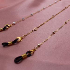 May include: Three gold eyeglass chains with different charms. The first chain has white pearls, the second has a heart charm, and the third has small gold beads.