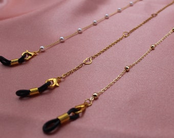 May include: Three gold eyeglass chains with different charms. The first chain has white pearls, the second has a heart charm, and the third has small gold beads.