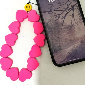 Puede incluir: Un charm de teléfono en forma de corazón rosa brillante con un charm de carita sonriente amarilla. El charm está junto a un smartphone negro con un fondo de pantalla de paisaje invernal.