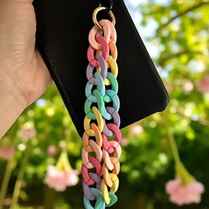 Puede incluir: Un adorno de teléfono colorido con un diseño de eslabones de cadena en tonos pastel de rosa, naranja, amarillo, verde y azul. El adorno está sujeto a una funda de teléfono negra con un anillo dorado.