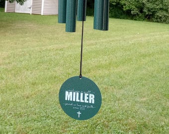 May include: A dark green wind chime featuring five cylindrical metal tubes suspended from a circular top. A round green wind sail hangs at the bottom with the text "melissa & scott MILLER joined in love and faith since 2003" and a cross symbol.