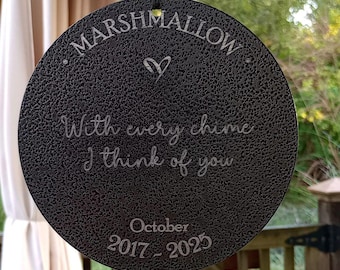 May include: A circular black textured wind chime plaque featuring the engraved text MARSHMALLOW, a heart symbol, the phrase With every chime I think of you, and the dates October 2017 - 2025, suspended by a black cord from a metal wind chime structure.