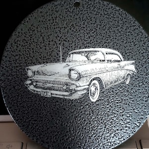 May include: A circular black metal ornament with a textured, hammered finish featuring a detailed white laser-etched illustration of a classic 1957 Chevrolet Bel Air car. The design includes a small hole at the top for hanging purposes.