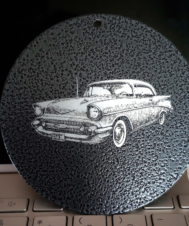 May include: A circular black metal ornament with a textured, hammered finish featuring a detailed white laser-etched illustration of a classic 1957 Chevrolet Bel Air car. The design includes a small hole at the top for hanging purposes.