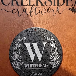 May include: A circular black textured sign featuring a white engraved letter W in the center, flanked by two leafy branches. Below the letter is the text WHITEHEAD and Est 2011. A small hole is at the top. The text CREEKSIDE craftwork is above.