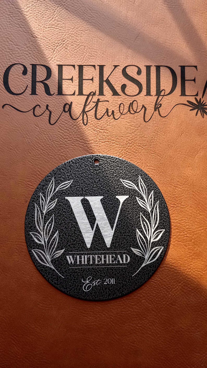 May include: A circular black textured sign featuring a white engraved letter W in the center, flanked by two leafy branches. Below the letter is the text WHITEHEAD and Est 2011. A small hole is at the top. The text CREEKSIDE craftwork is above.