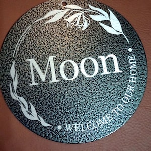 May include: A circular metal sign with a dark, textured, hammered finish. The center features the word "Moon" in large white letters, with the phrase "WELCOME TO OUR HOME" curved along the bottom. A white line drawing of a leafy vine encircles the text.