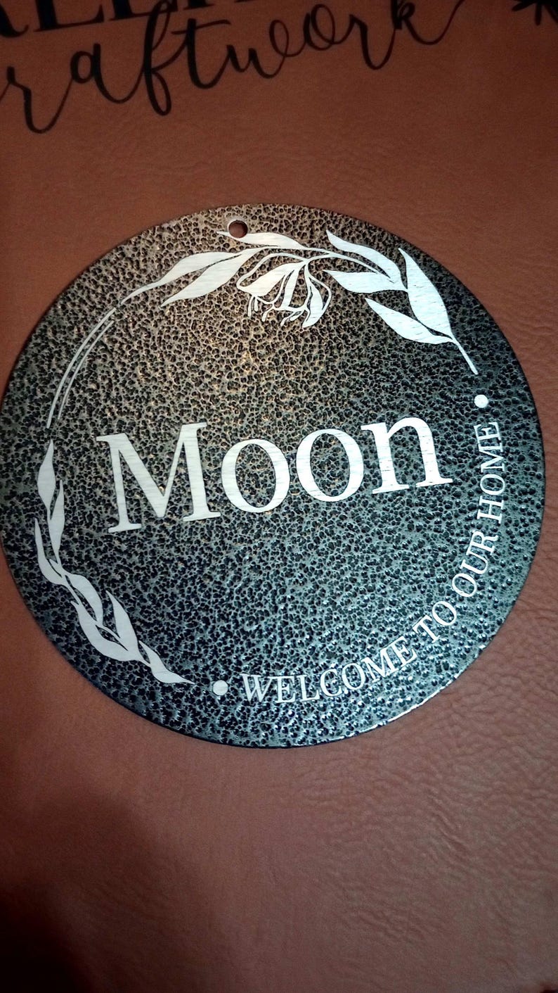 May include: A circular metal sign with a dark, textured, hammered finish. The center features the word "Moon" in large white letters, with the phrase "WELCOME TO OUR HOME" curved along the bottom. A white line drawing of a leafy vine encircles the text.