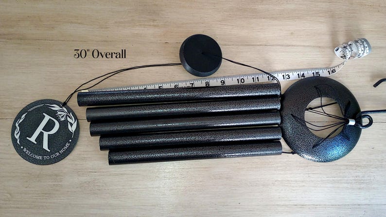 May include: A black wind chime with five cylindrical tubes and a circular top piece. A round hanging plaque features the letter R and the text Welcome to Our Home. A measuring tape is visible, and text indicates 30 inches overall length.