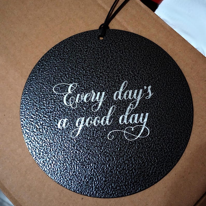May include: A circular black metal wall hanging with a textured finish. The text "Every day's a good day" is engraved in a white script font in the center. A thin black cord is attached to the top for hanging purposes.