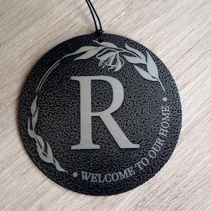 May include: A circular black sign with a textured finish featuring a large silver letter R in the center. A silver leaf wreath design surrounds the letter, and the text WELCOME TO OUR HOME is printed in silver along the bottom edge with a black string.