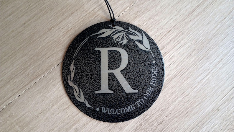 May include: A circular black sign with a textured finish featuring a large silver letter R in the center. A silver leaf wreath design surrounds the letter, and the text WELCOME TO OUR HOME is printed in silver along the bottom edge with a black string.