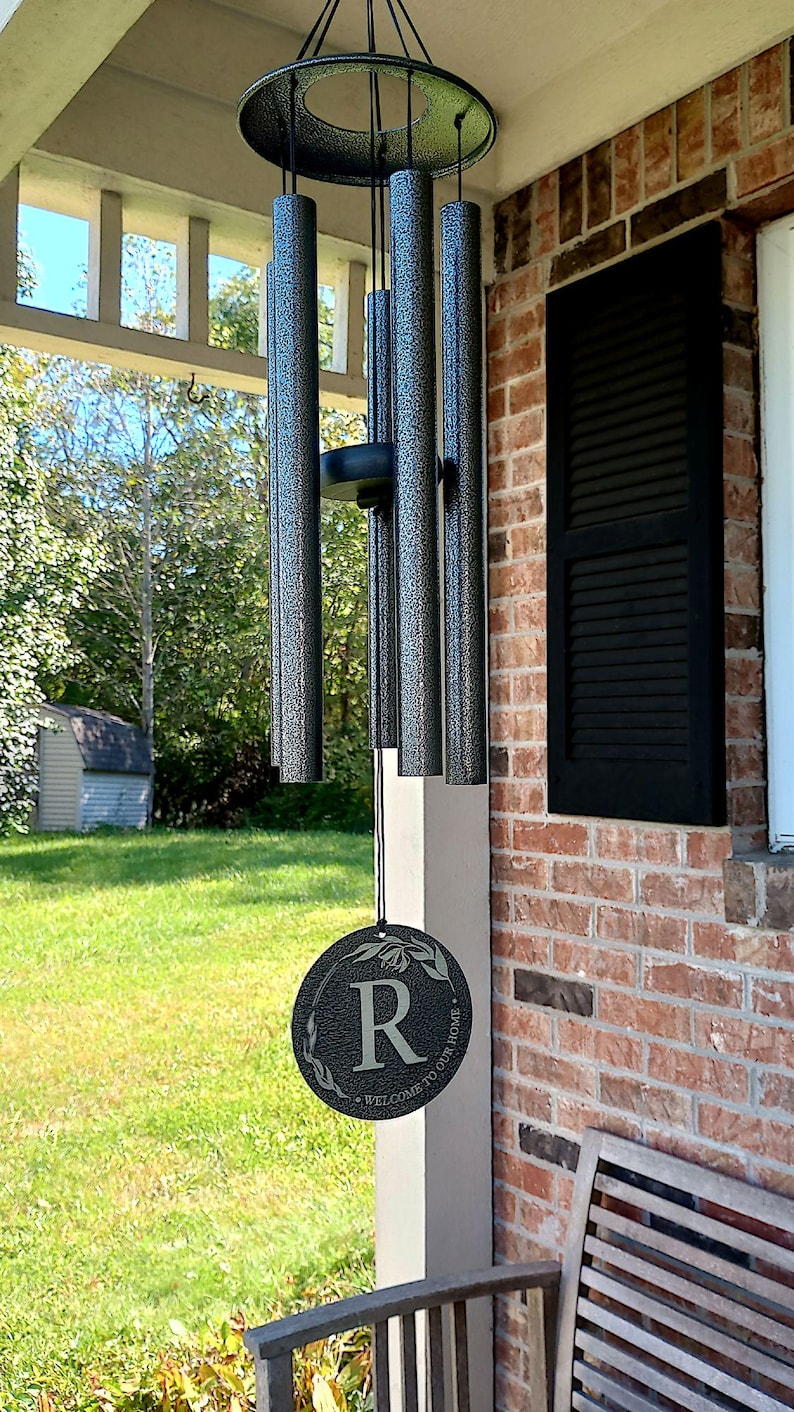 May include: A wind chime with six cylindrical tubes featuring a dark gray textured finish. The chime includes a circular wind catcher at the bottom, engraved with the letter R, a floral design, and the text Welcome to our home in white lettering.