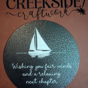 May include: A circular black metal plaque with a textured finish featuring a white graphic of a sailboat on water. Below the boat, the text reads: Wishing you fair winds and a relaxing next chapter. A small hole is at the top for hanging.