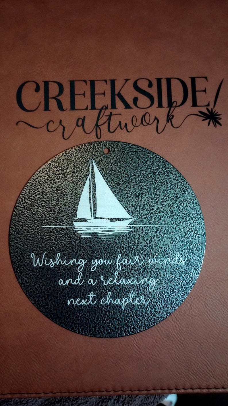 May include: A circular black metal plaque with a textured finish featuring a white graphic of a sailboat on water. Below the boat, the text reads: Wishing you fair winds and a relaxing next chapter. A small hole is at the top for hanging.