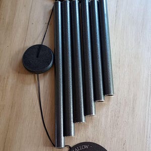 May include: A dark gray metal wind chime featuring five cylindrical tubes, a circular top canopy, and a round wind catcher. The wind catcher is engraved with the text "MARSHMALLOW", "With every chime I think of you", and "October 2017 - 2025".