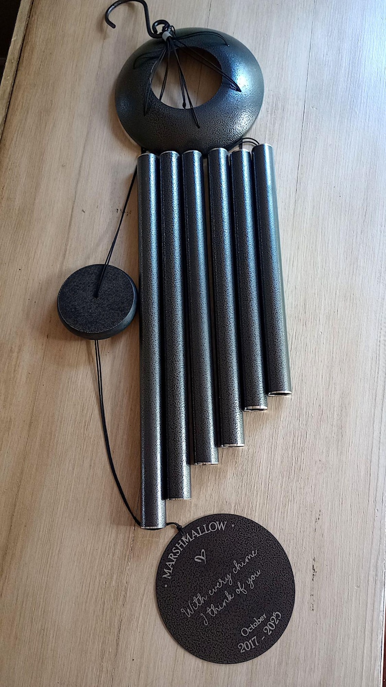May include: A dark gray metal wind chime featuring five cylindrical tubes, a circular top canopy, and a round wind catcher. The wind catcher is engraved with the text "MARSHMALLOW", "With every chime I think of you", and "October 2017 - 2025".