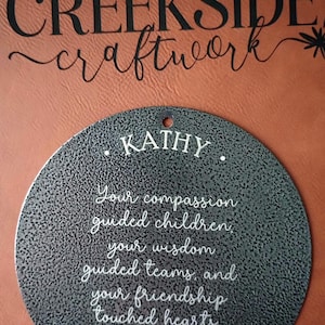 May include: A circular dark gray metal plaque with a textured finish featuring the white engraved text: KATHY. Your compassion guided children, your wisdom guided teams, and your friendship touched hearts. A small white heart icon is at the bottom.