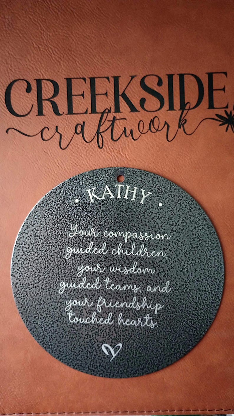 May include: A circular dark gray metal plaque with a textured finish featuring the white engraved text: KATHY. Your compassion guided children, your wisdom guided teams, and your friendship touched hearts. A small white heart icon is at the bottom.