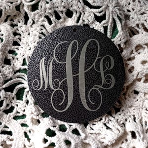 May include: A circular black ornament with a textured surface featuring a silver-toned engraved monogram with the letters M, H, and L in a decorative script font. The ornament has a small hole at the top for hanging and a smooth metallic finish.