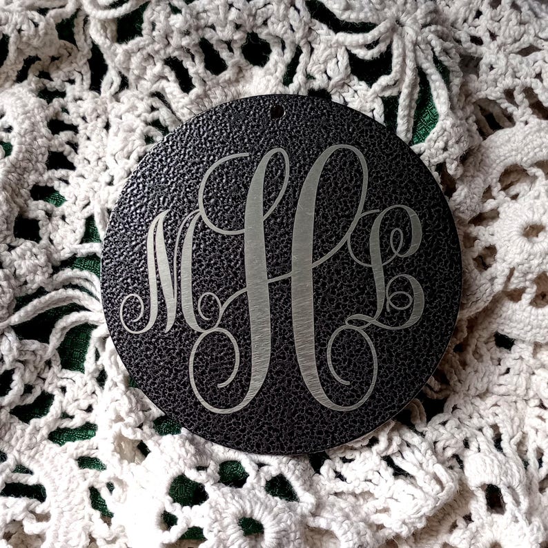 May include: A circular black ornament with a textured surface featuring a silver-toned engraved monogram with the letters M, H, and L in a decorative script font. The ornament has a small hole at the top for hanging and a smooth metallic finish.