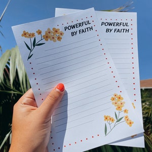 May include: Two white lined notepads with red dotted borders and yellow flower illustrations. The top of each notepad says "Powerful by Faith".