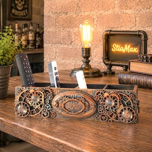 Puede incluir: Un organizador de escritorio rectangular con acabado en tono cobre y engranajes decorativos. La placa ovalada central muestra el texto, THE EYE IS THE MIRROR OF THE SOUL. Contiene tres mandos a distancia en sus compartimentos.