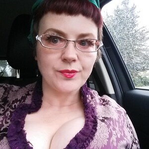 Peut inclure: Une personne portant des lunettes et une robe &agrave; motifs floraux violets, assise dans une voiture. La personne a du rouge &agrave; l&egrave;vres rouge et un foulard vert. La fen&ecirc;tre de la voiture montre une sc&egrave;ne ext&eacute;rieure pluvieuse.