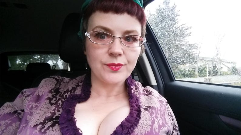 Peut inclure: Une personne portant des lunettes et une robe &agrave; motifs floraux violets, assise dans une voiture. La personne a du rouge &agrave; l&egrave;vres rouge et un foulard vert. La fen&ecirc;tre de la voiture montre une sc&egrave;ne ext&eacute;rieure pluvieuse.